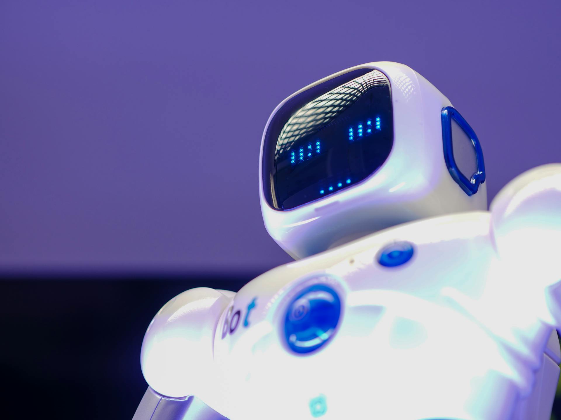 Close-up of a small white humanoid robot with a digital face illuminated in blue light against a purple background.